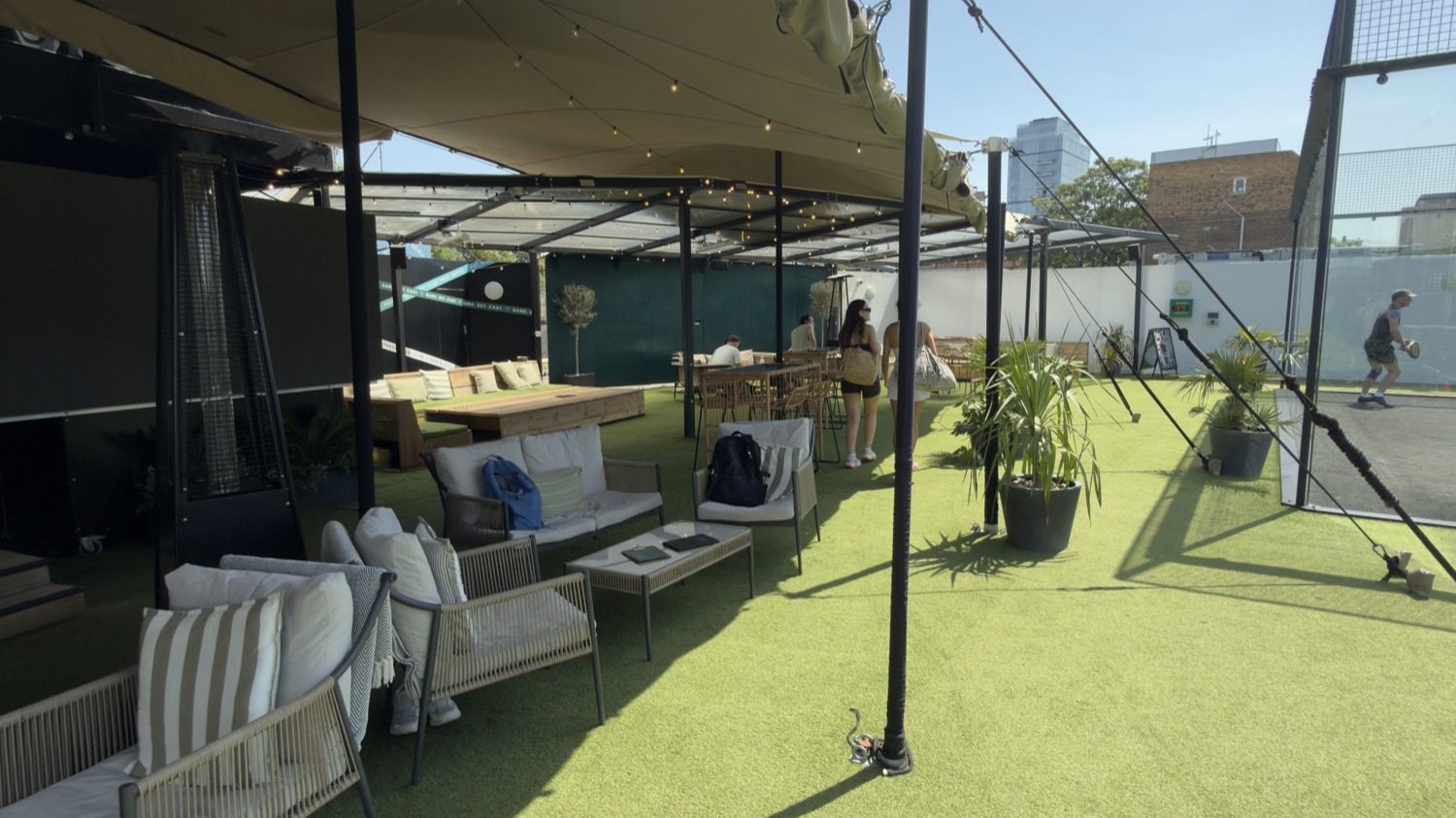 The fairy-lit canopy, bar and turf at Padel Social Club Earls Court, with loungers and players across the club.