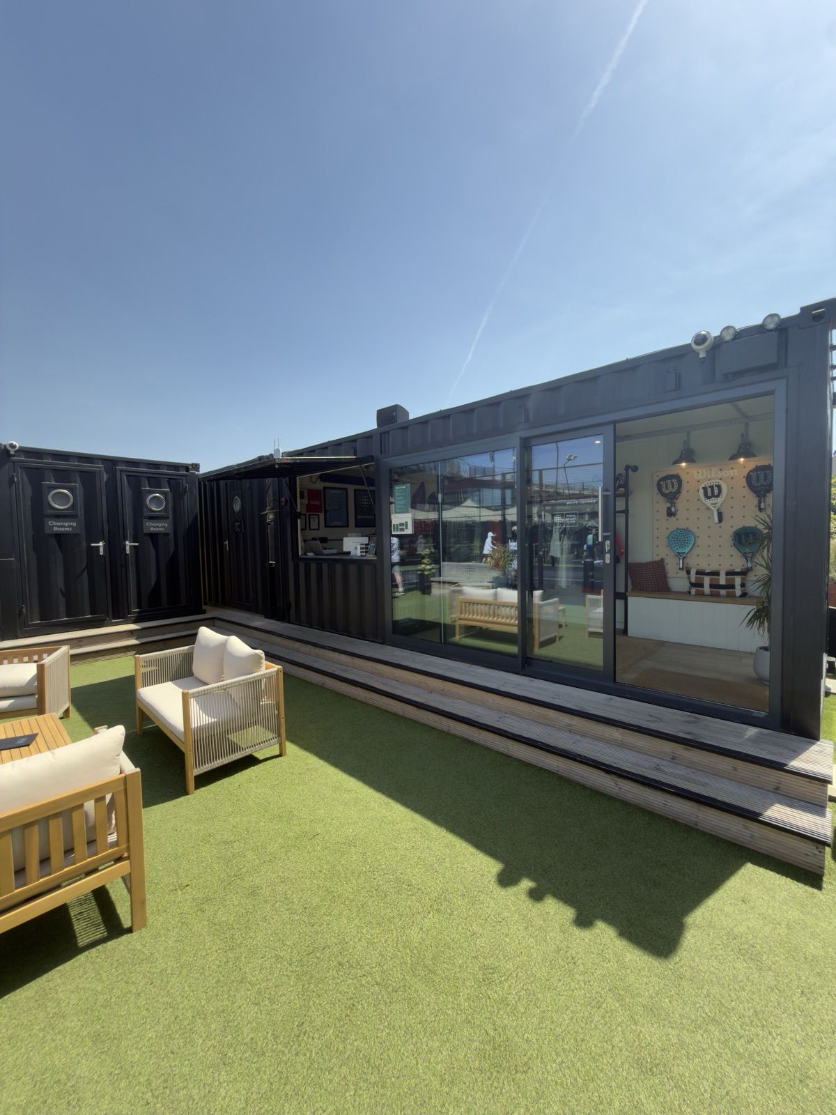 The black shipping-container changing rooms and pro shop at Padel Social Club Earls Court, with lounge seating on the turf.